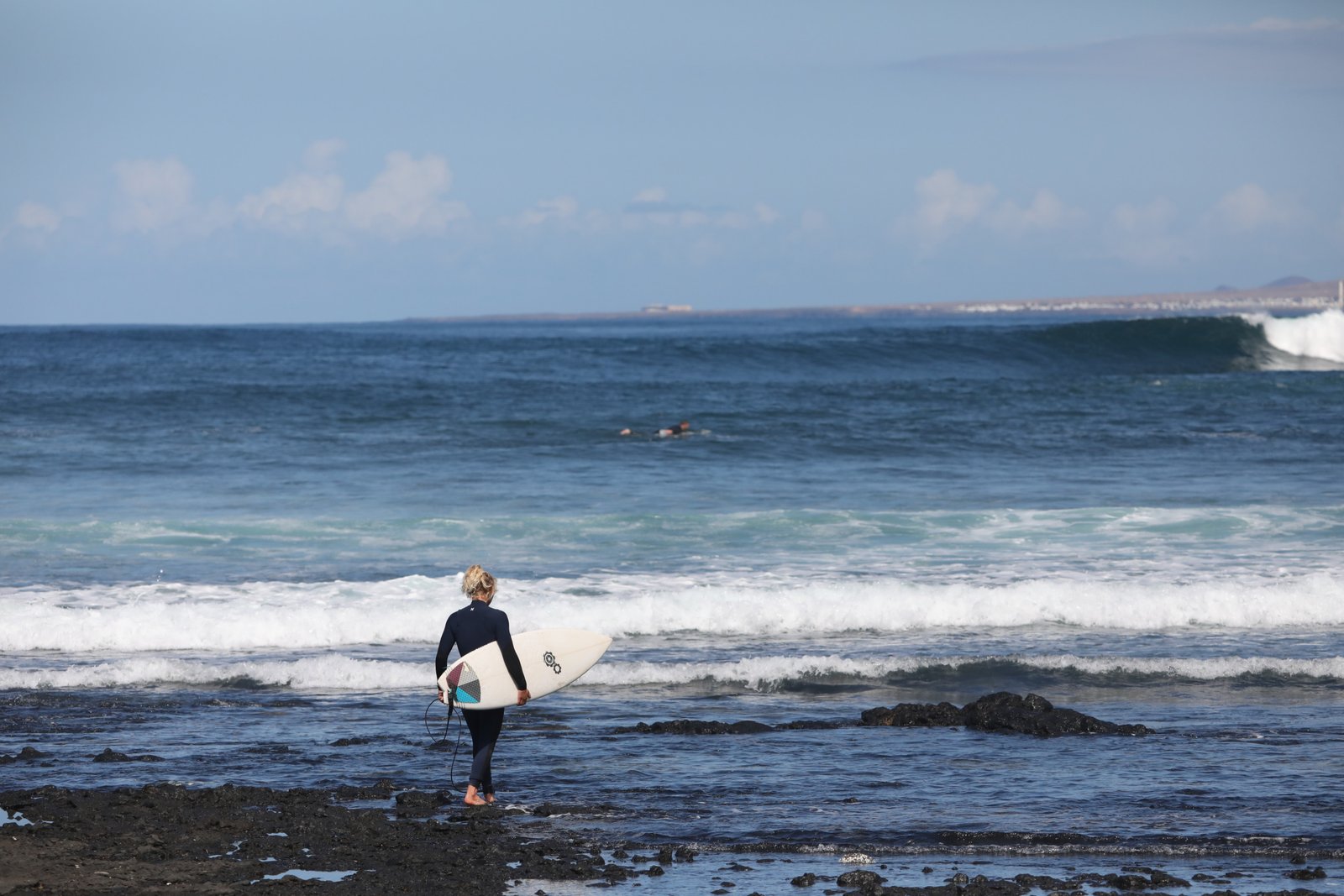 A surfer in a dark wetsuit stands on a rocky shoreline in Fuerteventura holding a white surfboard, gazing out at breaking ocean waves during surf lessons, with a distant Canary Islands coastline and partly cloudy sky on the horizon