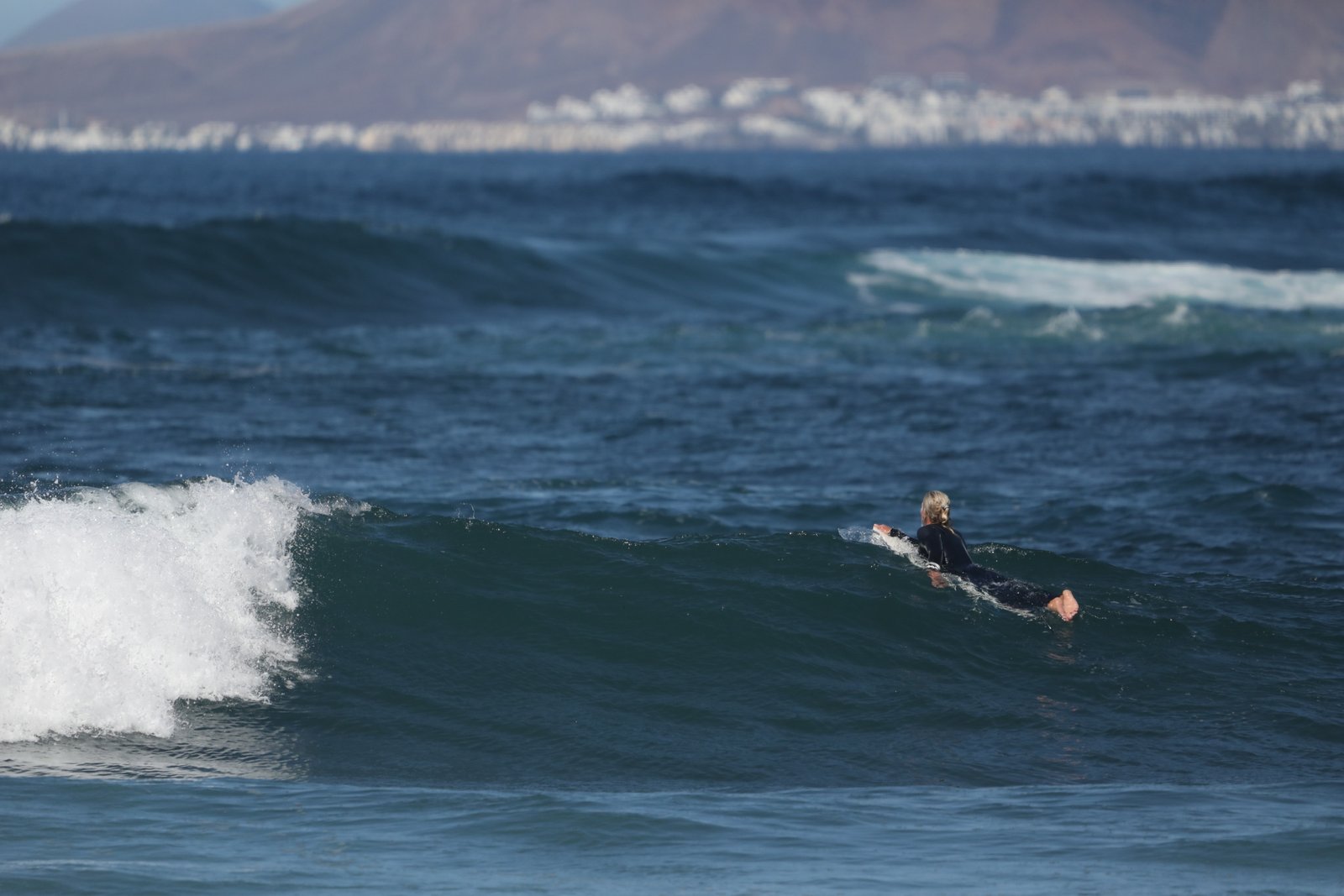 A surfer in a dark wetsuit paddles through a breaking ocean wave during surf lessons in Fuerteventura, with the arid Canary Islands mountains and coastline visible in the background.