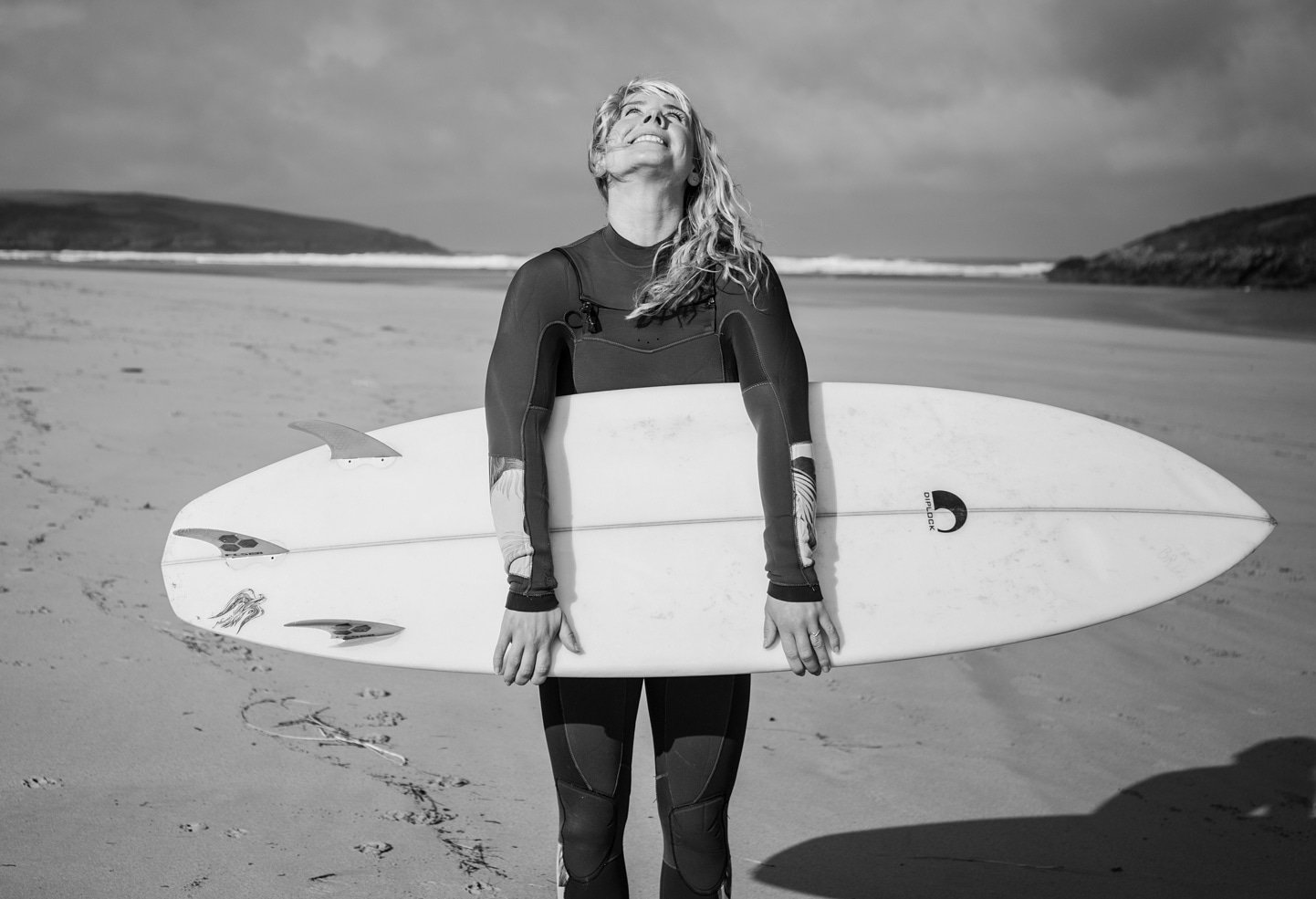 Black and white portrait of Michalina, surf coach and ISA World Championship competitor, laughing with her head tilted back while holding a Dharlock surfboard on a windswept beach.
