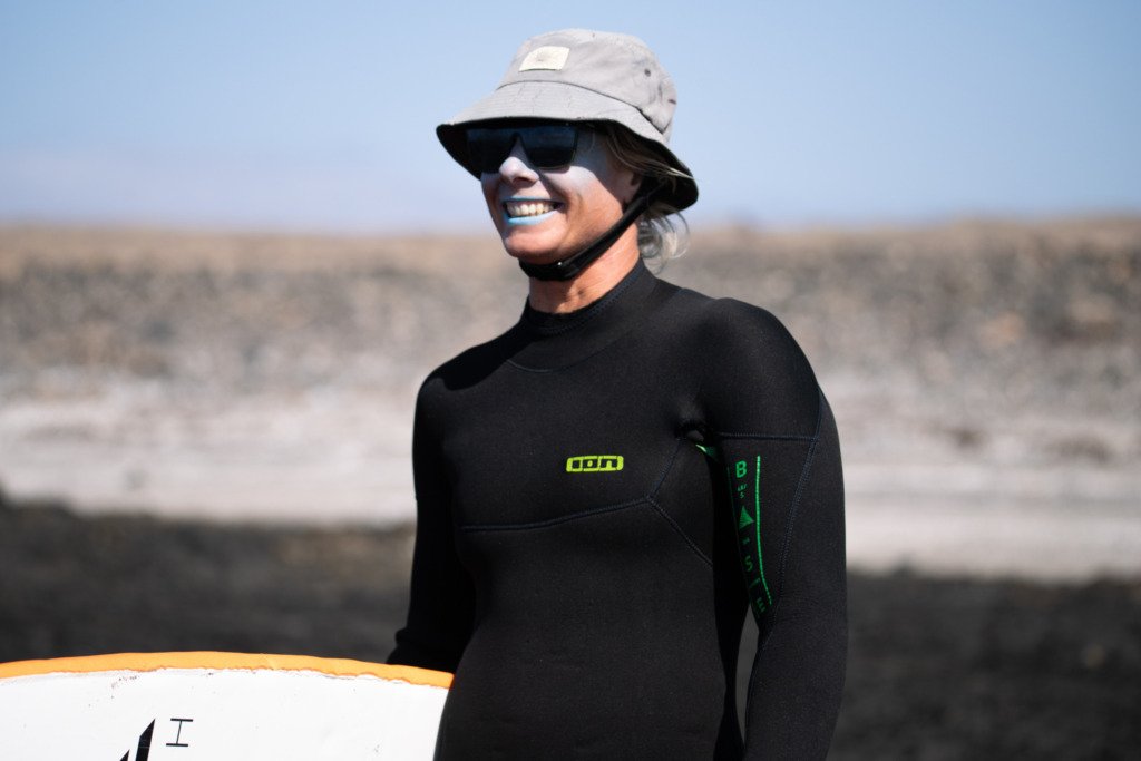 Michalina, private surf coach based in Corralejo, Fuerteventura, smiling in a black ION wetsuit, bucket hat, and sunglasses, holding a surfboard against an arid volcanic landscape backdrop.