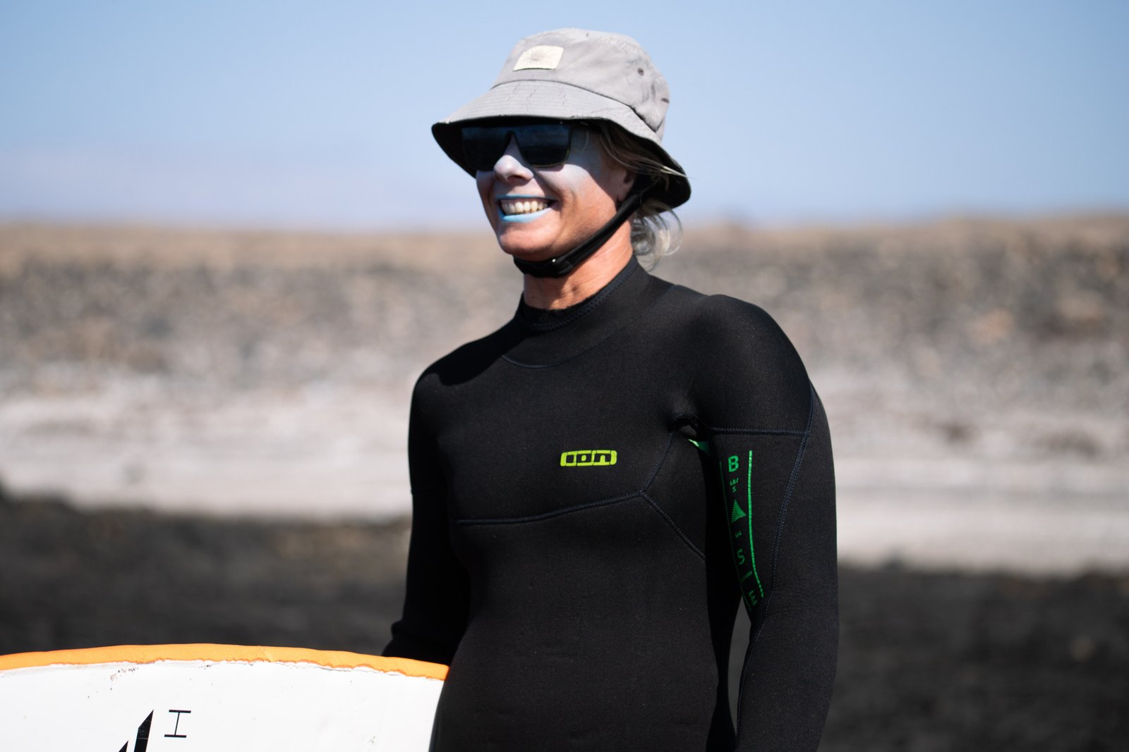 Michalina, private surf coach based in Corralejo, Fuerteventura, smiling in a black ION wetsuit, bucket hat, and sunglasses, holding a surfboard against an arid volcanic landscape backdrop.