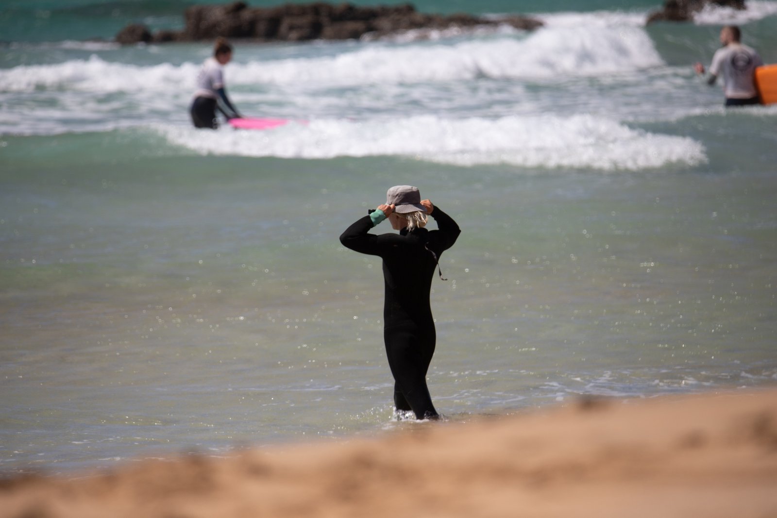 Michalina, surf coach in Fuerteventura, standing waist-deep in the ocean adjusting her bucket hat while watching over students during a surf lesson in Corralejo, with surfers practising on boards visible in the breaking waves behind her.