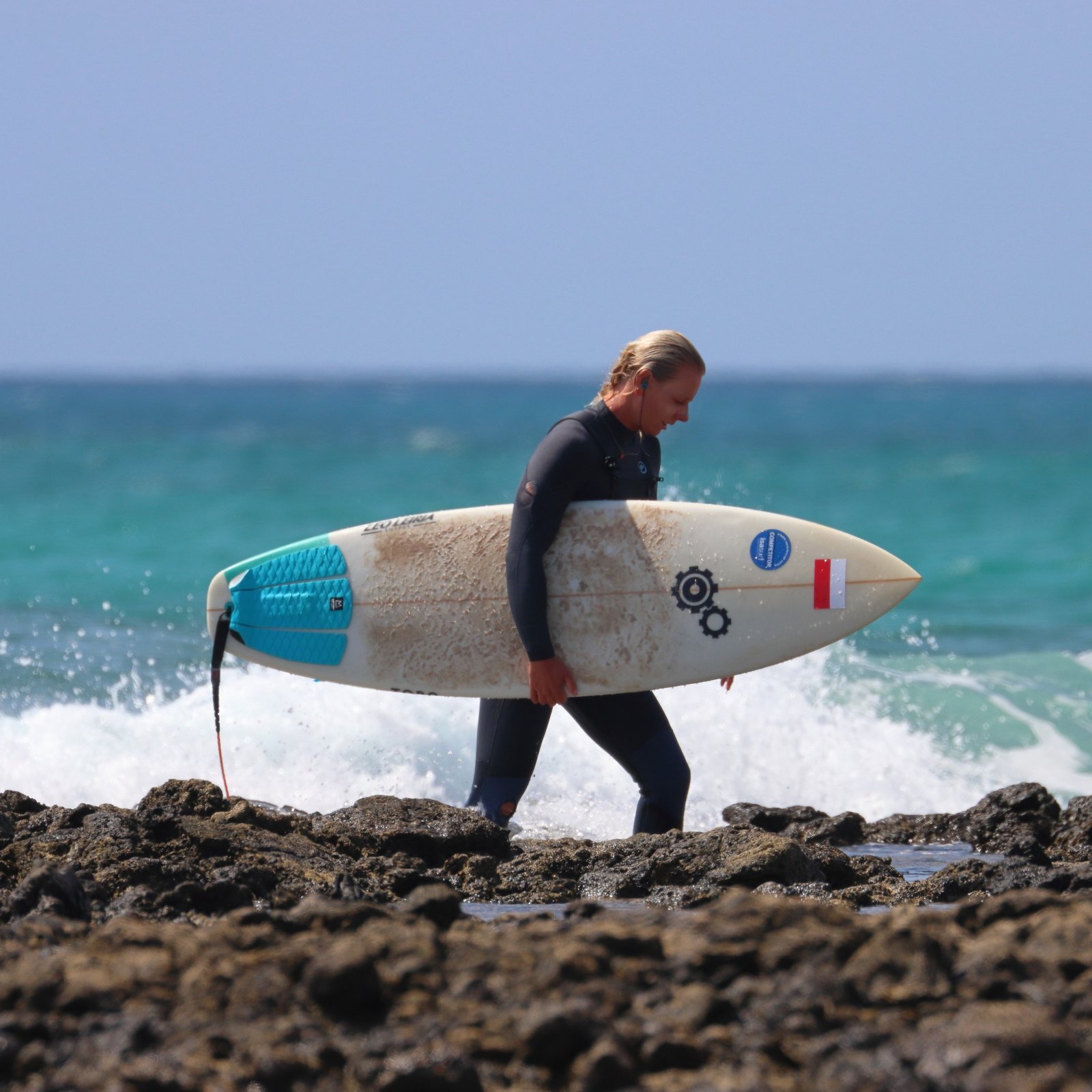 Michalina, private surf coach in Fuerteventura, walking across volcanic rocks carrying a waxed shortboard after a surf session in Corralejo, with turquoise Atlantic waves breaking behind her.